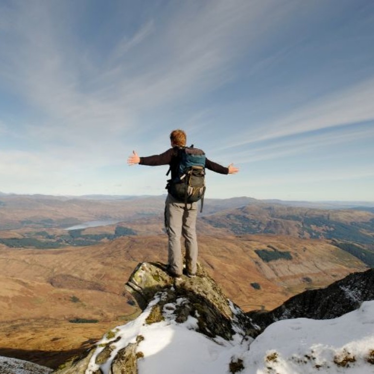 Person with a backpack stands on a rocky peak with arms outstretched, overlooking a vast mountainous landscape.