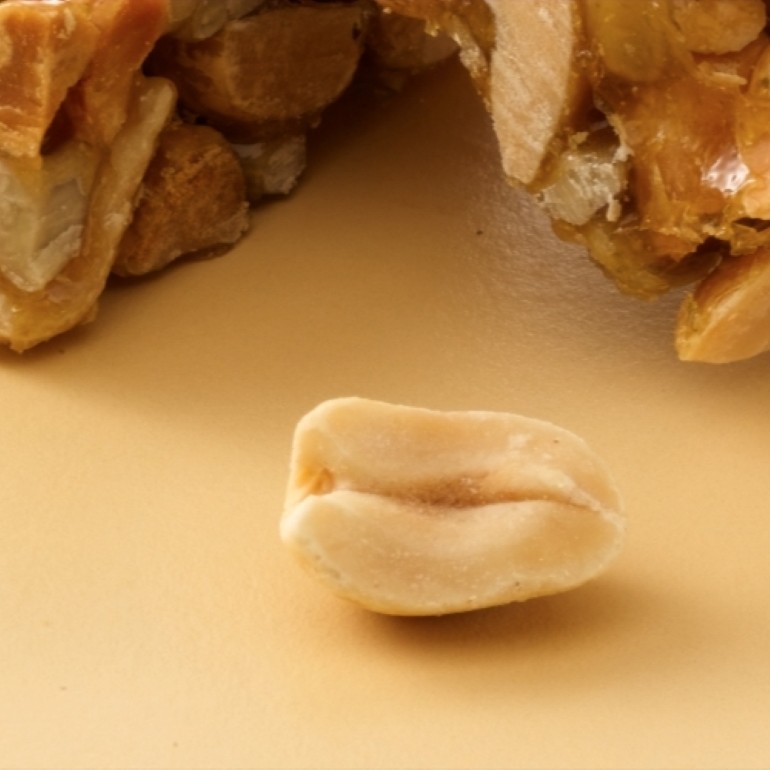 Single peanut kernel in focus on a beige surface, with caramel-coated peanut clusters softly blurred in the background.