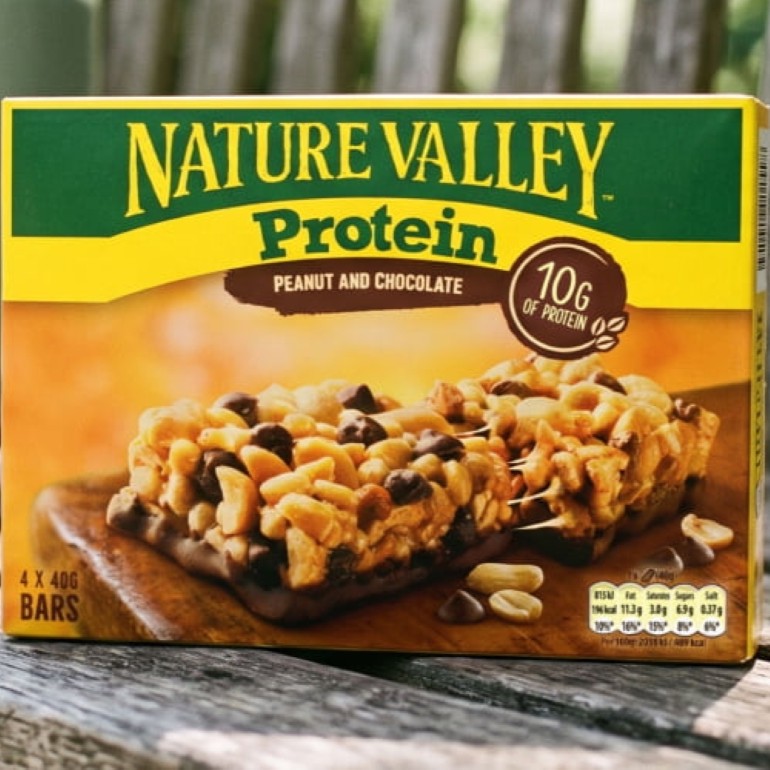 Box of Nature Valley Protein Peanut & Chocolate snack bars displayed on a wooden surface. The green and yellow packaging features two peanut and chocolate bars, highlights 10g of protein, and indicates a pack of 4 x 40g bars.