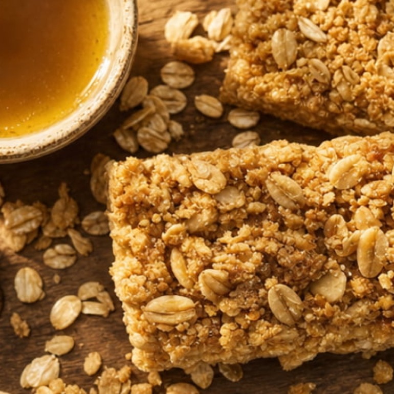 Homemade oat snack bars on a wooden surface, topped with rolled oats and crumbs, alongside a bowl of golden honey.