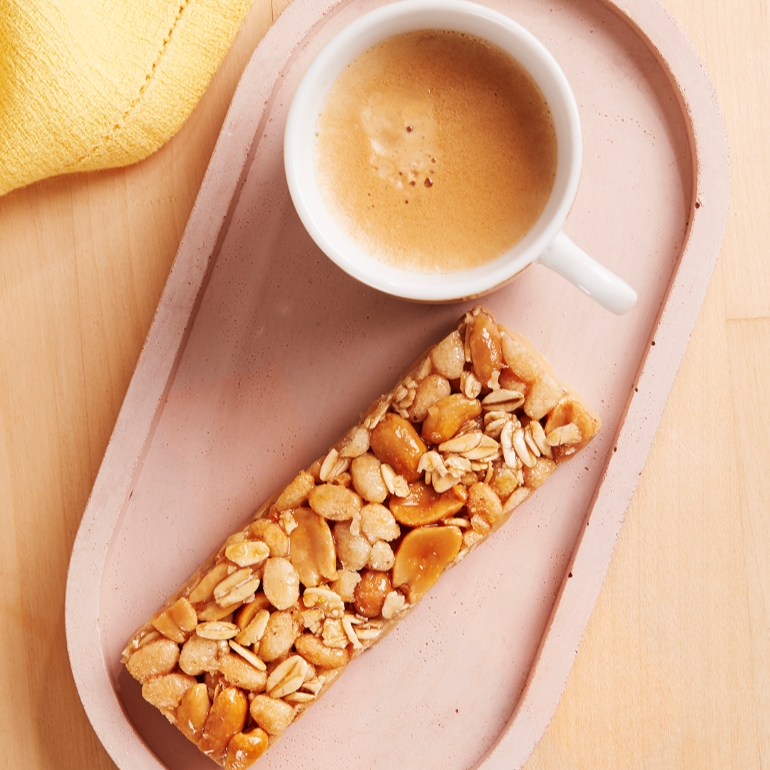 A pink tray holds a cup of coffee and a granola bar topped with oats and nuts.