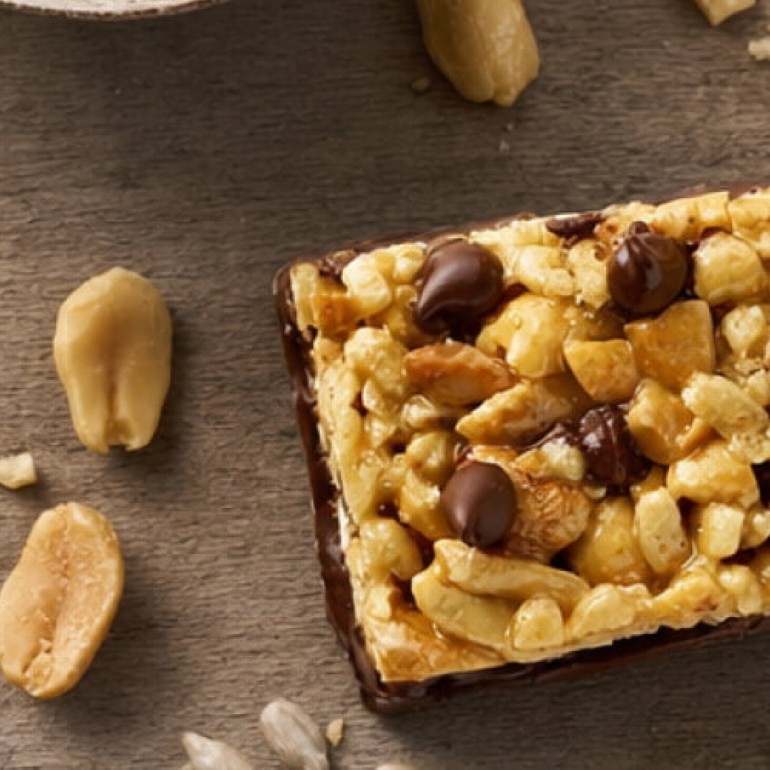 Close-up of a nut and chocolate snack bar topped with chopped peanuts, cashews, and chocolate chips on a dark wooden surface, with loose peanuts scattered around the bar.