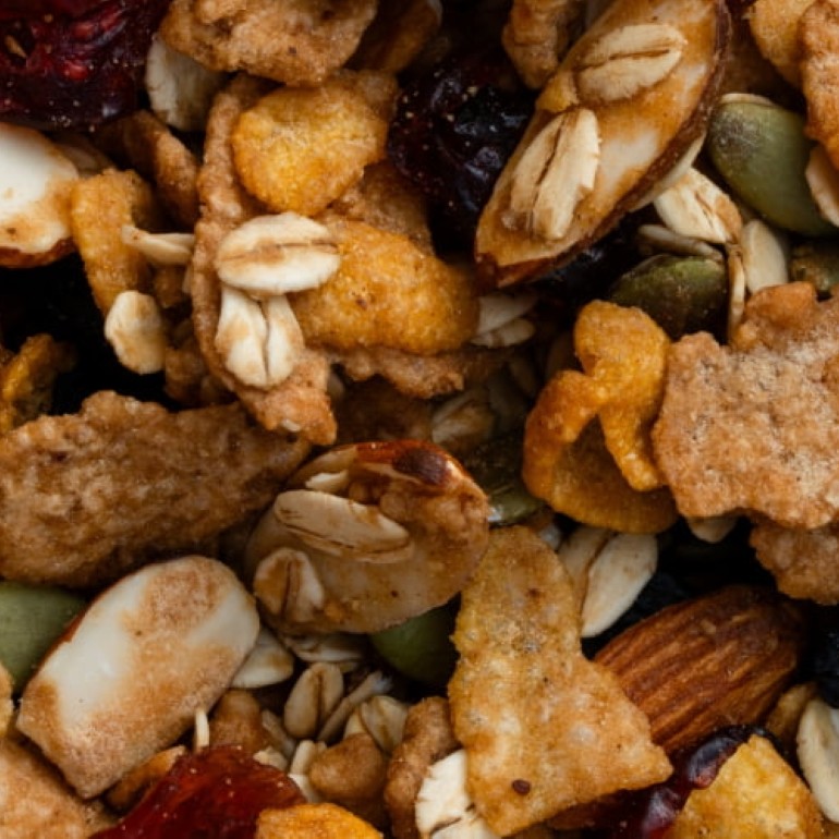 Close-up of a mixed granola and trail mix blend featuring rolled oats, almonds, peanuts, pumpkin seeds, dried cranberries, and crunchy cereal clusters in shades of golden brown, cream, green, and deep red.