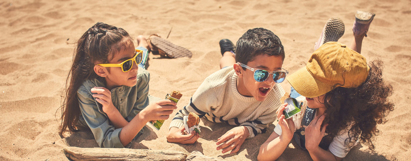 Three children lay in the sand eating Nature Valley bars