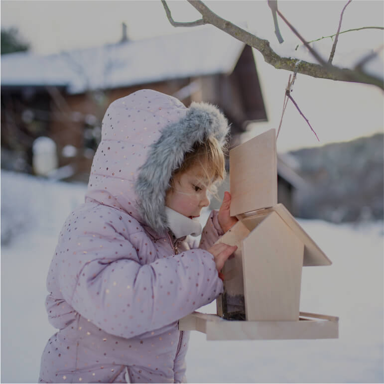 small girl wearing winter jacket opening bird feeder surrounded by snow