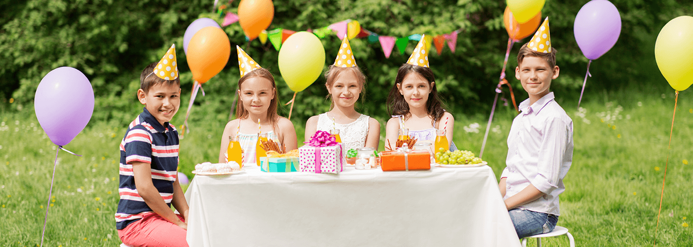 A group of five kids celebrating birthday party around a table placed with gifts and food in garden