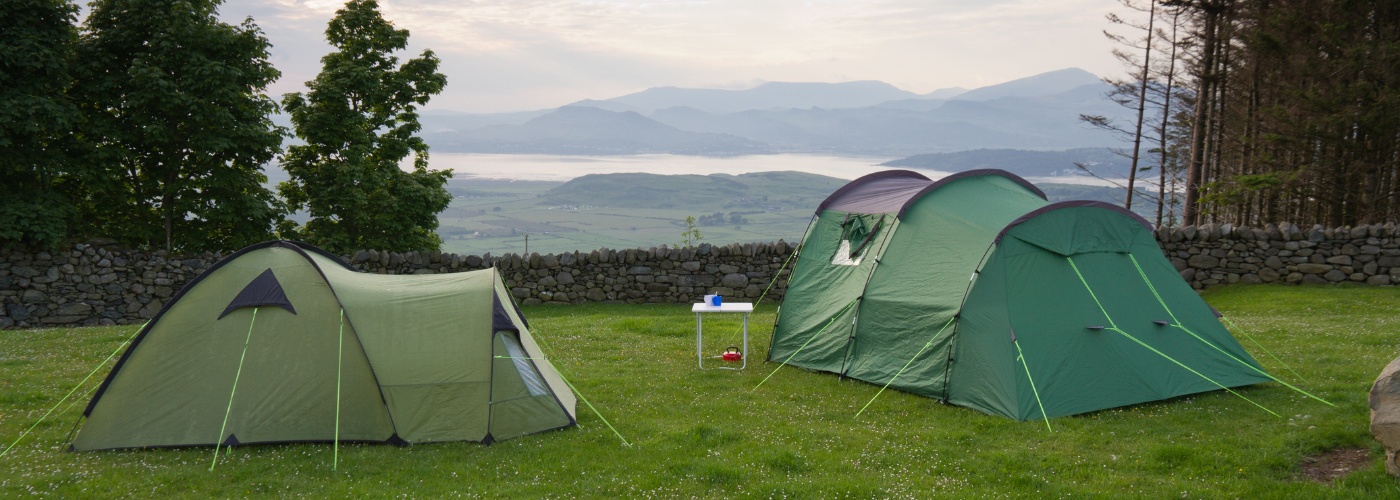 Two green coloured camping tents on grass facing a valley of lake and mountains.