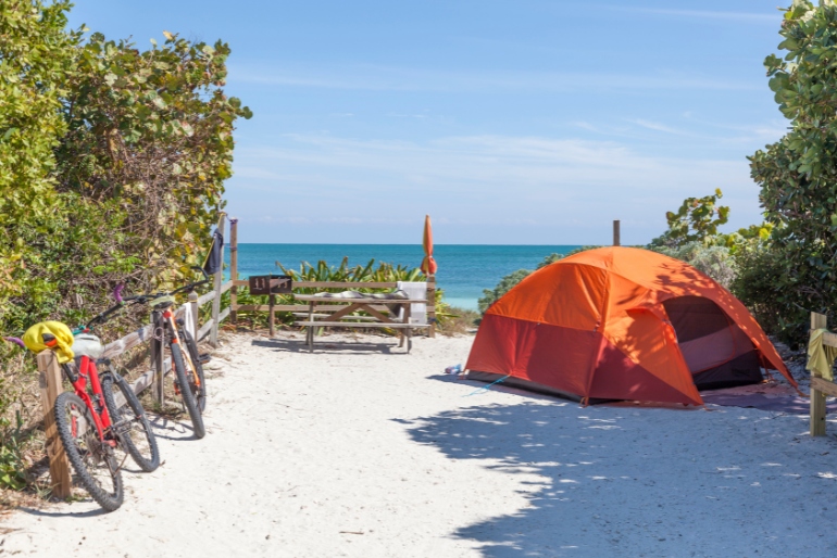 An orange camping tent on white sand facing the beach. Two cycles parked outside the tent.